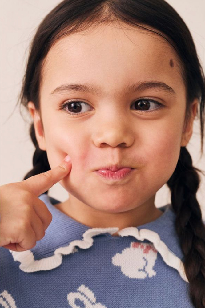 Close-up of a young girl with braided pigtails poking her cheek while wearing a blue knitted rabbit sweater.