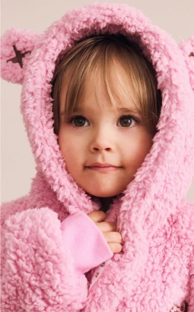 Young child wearing a textured pink fleece hoodie with teddy bear ears against a plain light backdrop.