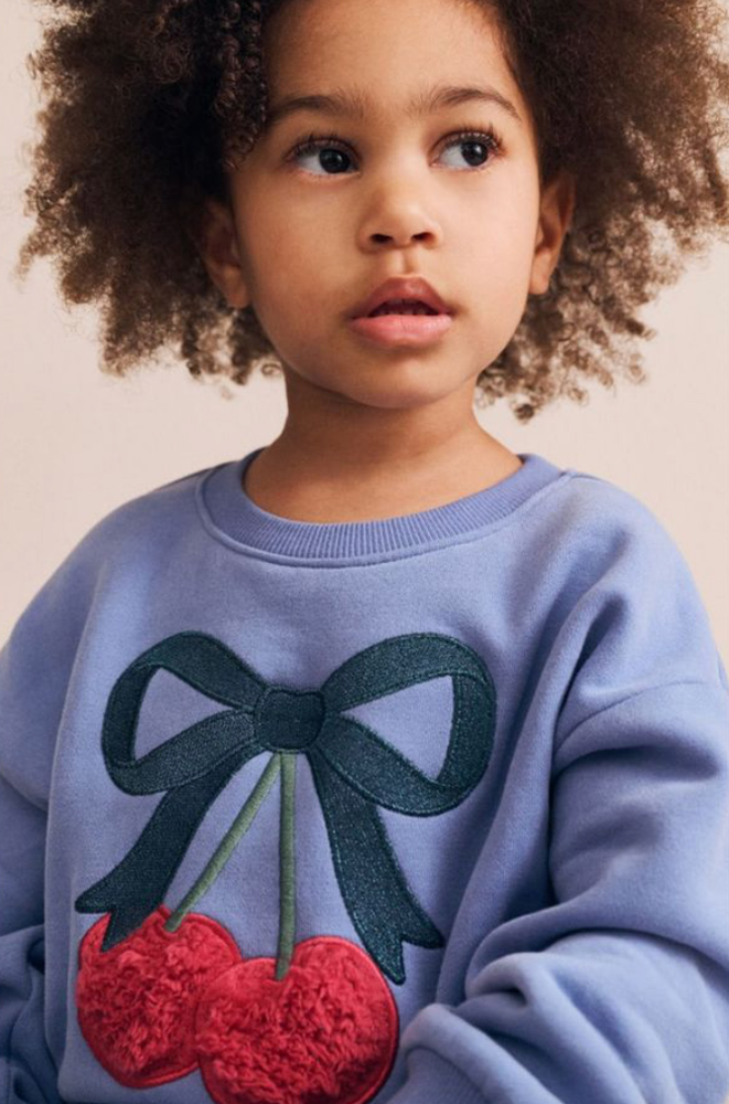 Young child with curly hair wearing a periwinkle sweatshirt featuring a cherry and bow appliqué against a plain neutral backdrop.