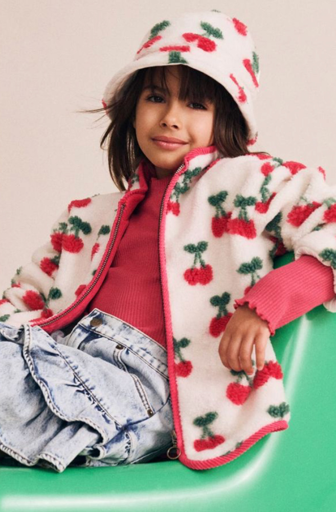 Girl wearing a white cherry-pattern fleece jacket, matching bucket hat, and denim skirt seated on a green chair.