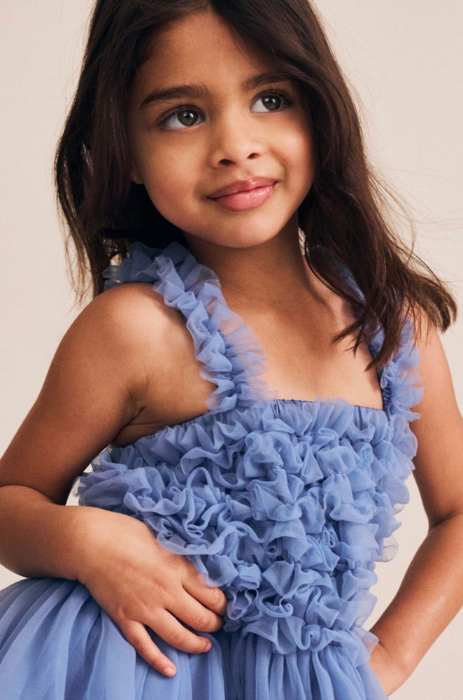 Young girl with long brown hair posing in a textured ruffled light blue tulle dress against a warm neutral studio backdrop.