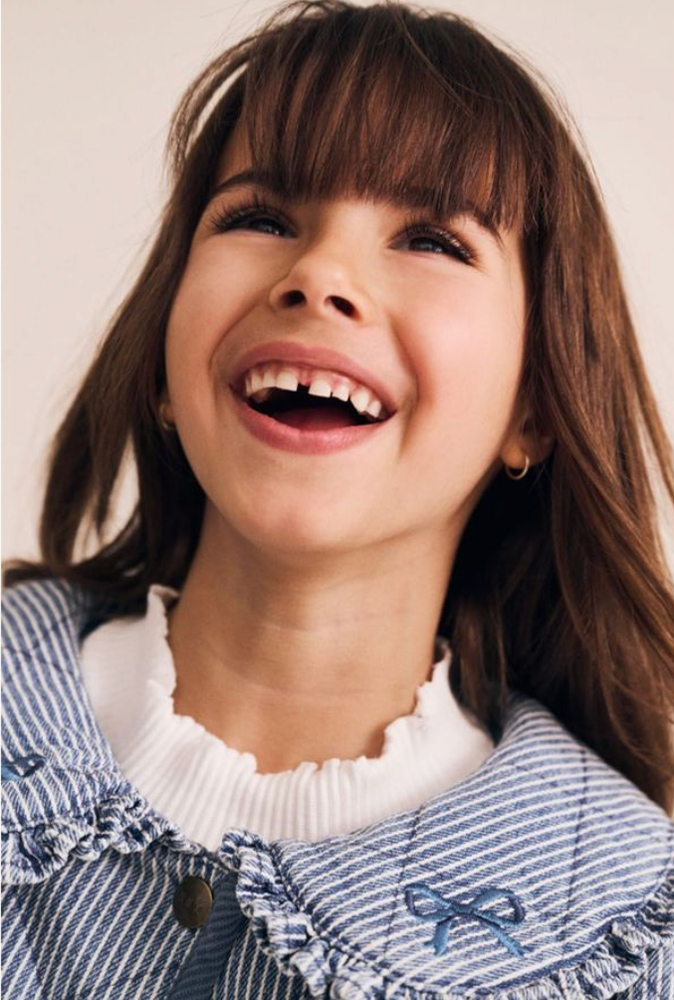 Close-up portrait of a laughing young girl with bangs wearing a blue striped quilted jacket with a ruffled collar.