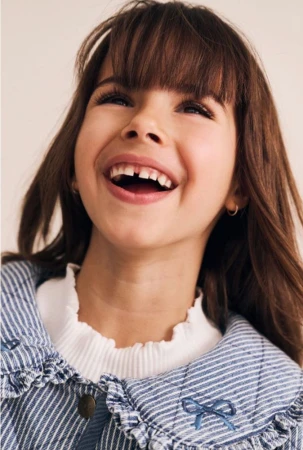 Close-up portrait of a laughing young girl with bangs wearing a blue striped quilted jacket with a ruffled collar.