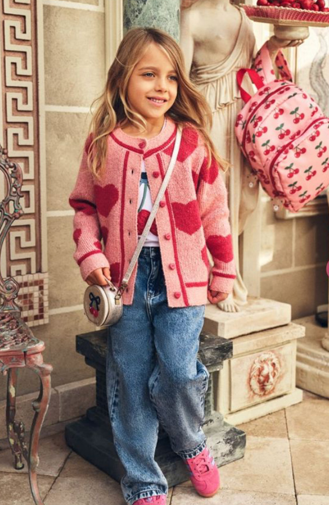 Young girl smiling in a pink knit cardigan with red hearts, wide blue denim jeans, and pink sneakers with a round crossbody bag.