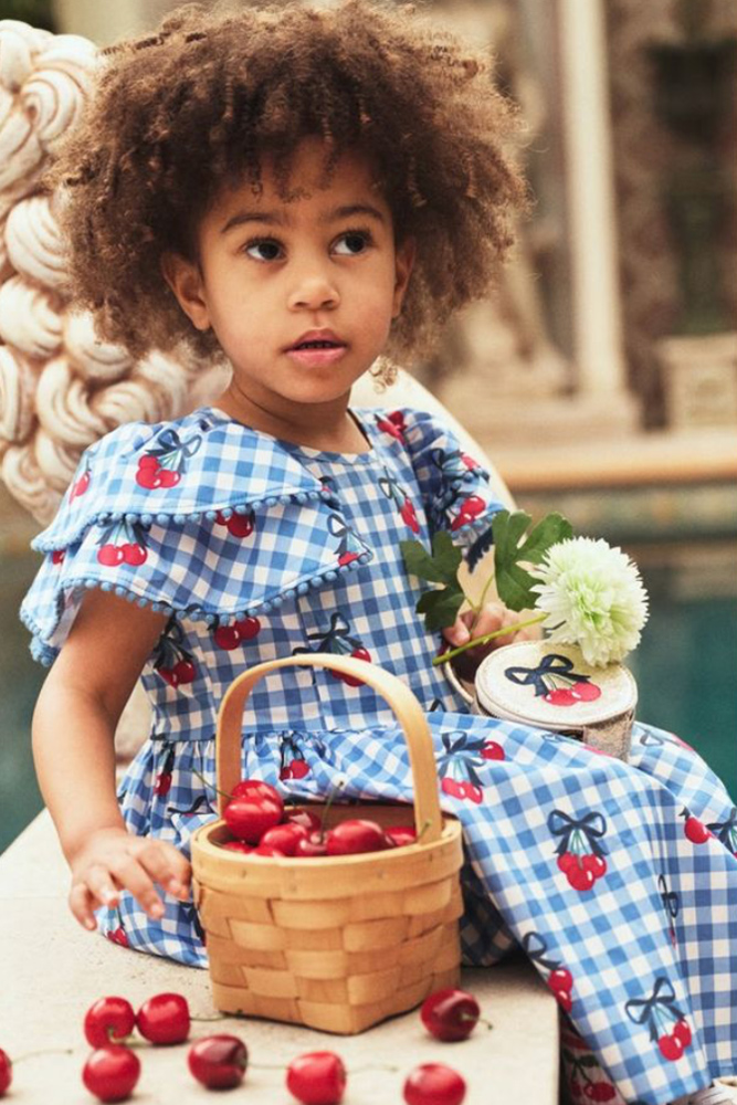 Young girl with curly hair in a blue gingham cherry print dress seated beside a small basket of fresh cherries outdoors.