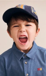 Young boy shouting with mouth open, wearing a blue denim collared shirt and baseball cap against a neutral backdrop.