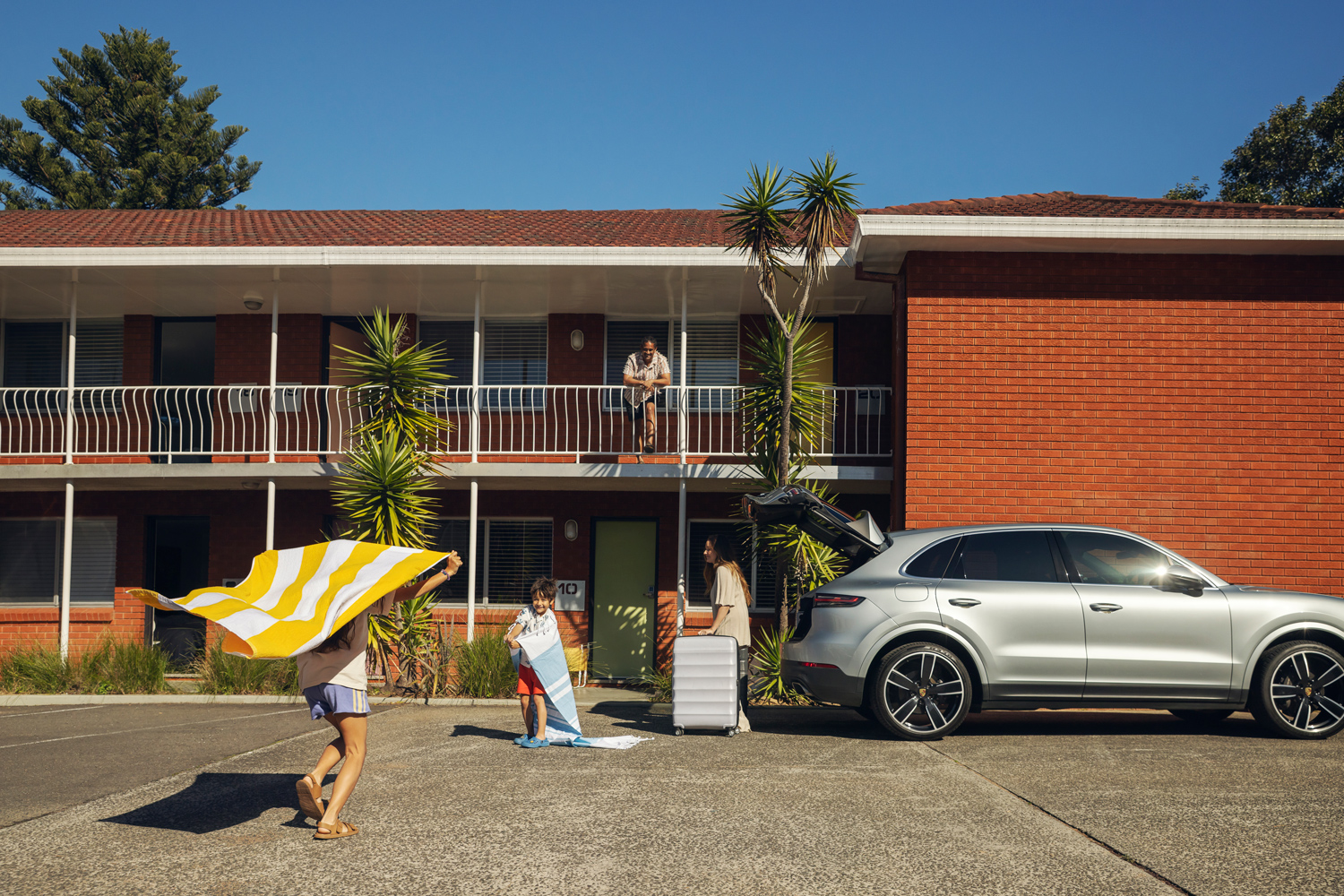 A family arrives at a brick motel with luggage and beach towels beside a parked silver SUV with an open trunk.