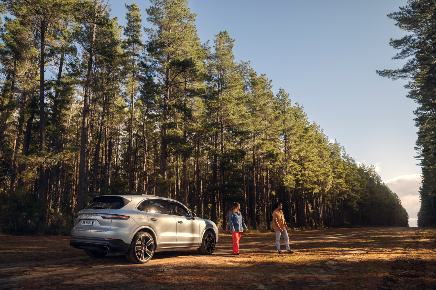 Silver luxury SUV parked on a dirt forest track in warm sunlight beside a man and woman standing near tall pine trees.
