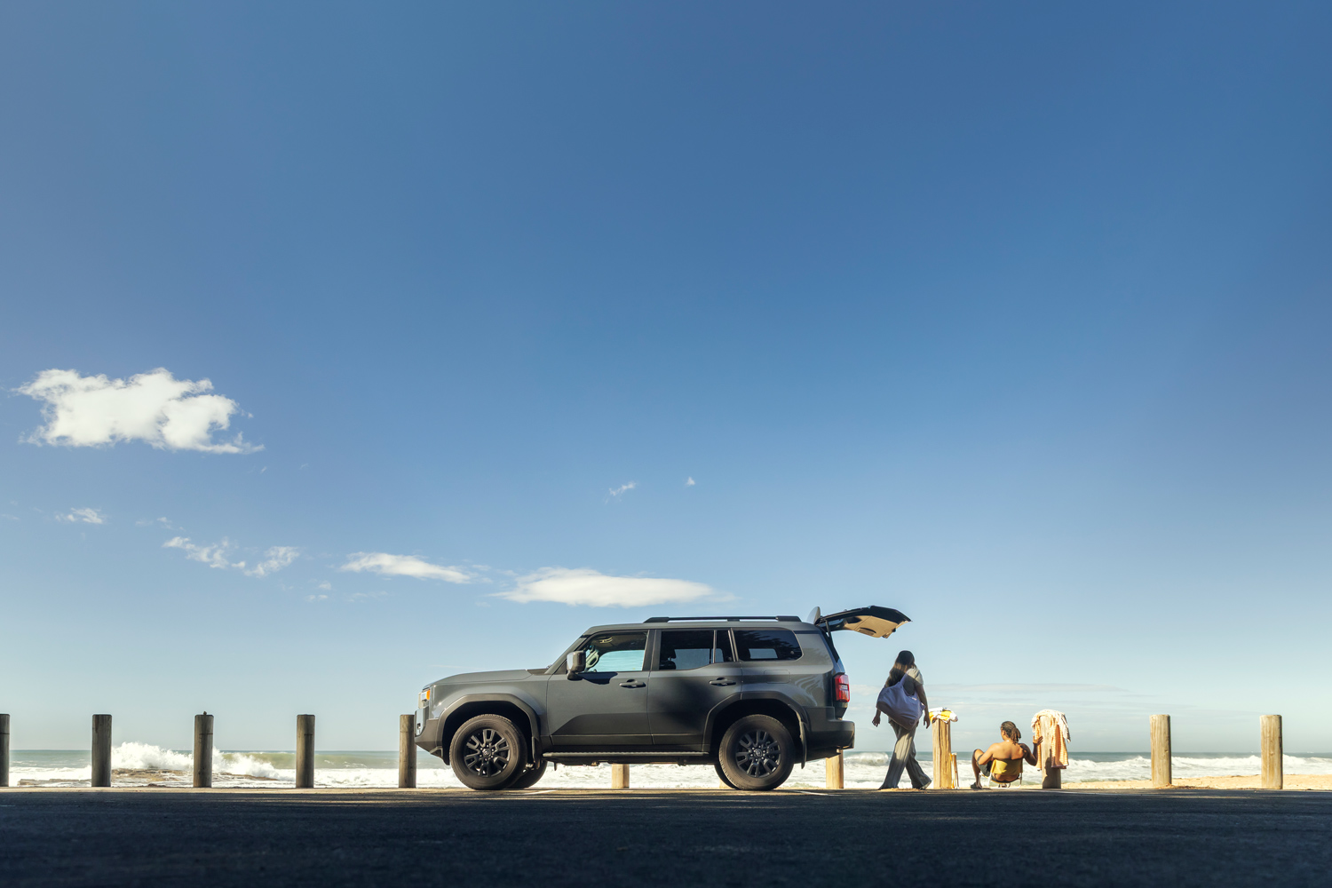 Dark grey SUV parked near a coastal beach barrier with open rear trunk beside two people under a wide clear blue sky.