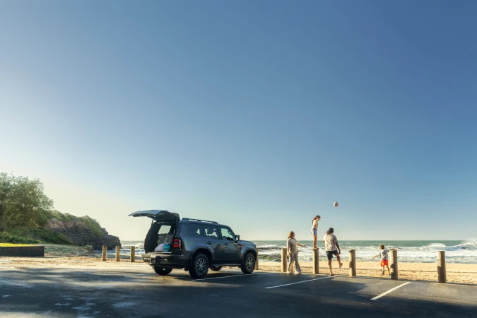 Family near a parked SUV with its hatch open at a coastal parking lot overlooking the ocean.