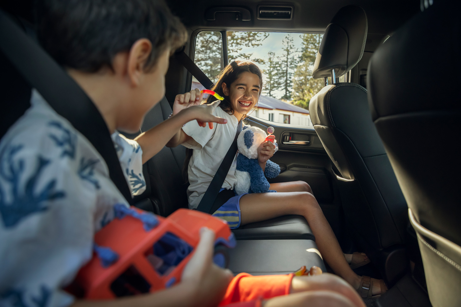 Two children smiling and playing with toys and candy in the back seat of a car during a road trip.