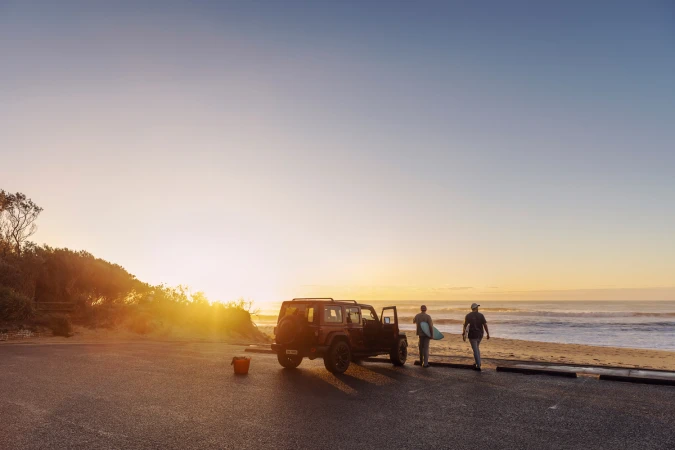 Two surfers carry surfboards from a parked 4x4 SUV toward the ocean at golden hour sunrise.
