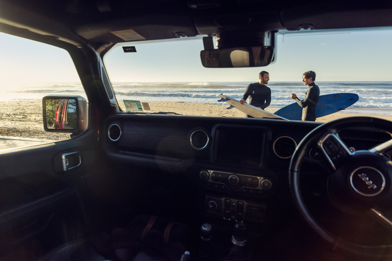 View from inside a car showing two surfers in wetsuits holding surfboards on a sunny beach by the ocean.