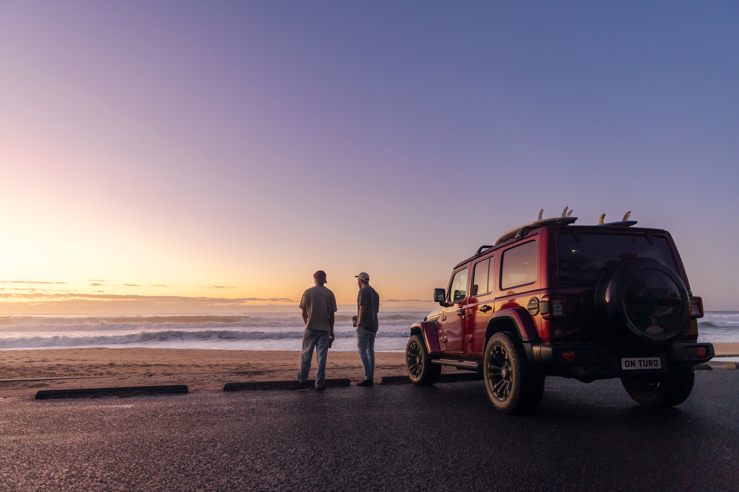 Two men standing by a red SUV with surfboards on a beach at sunset