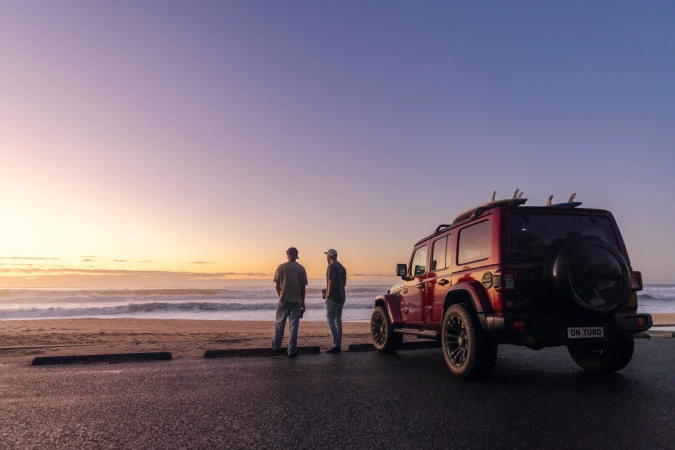Two men standing by a red SUV with surfboards on a beach at sunset