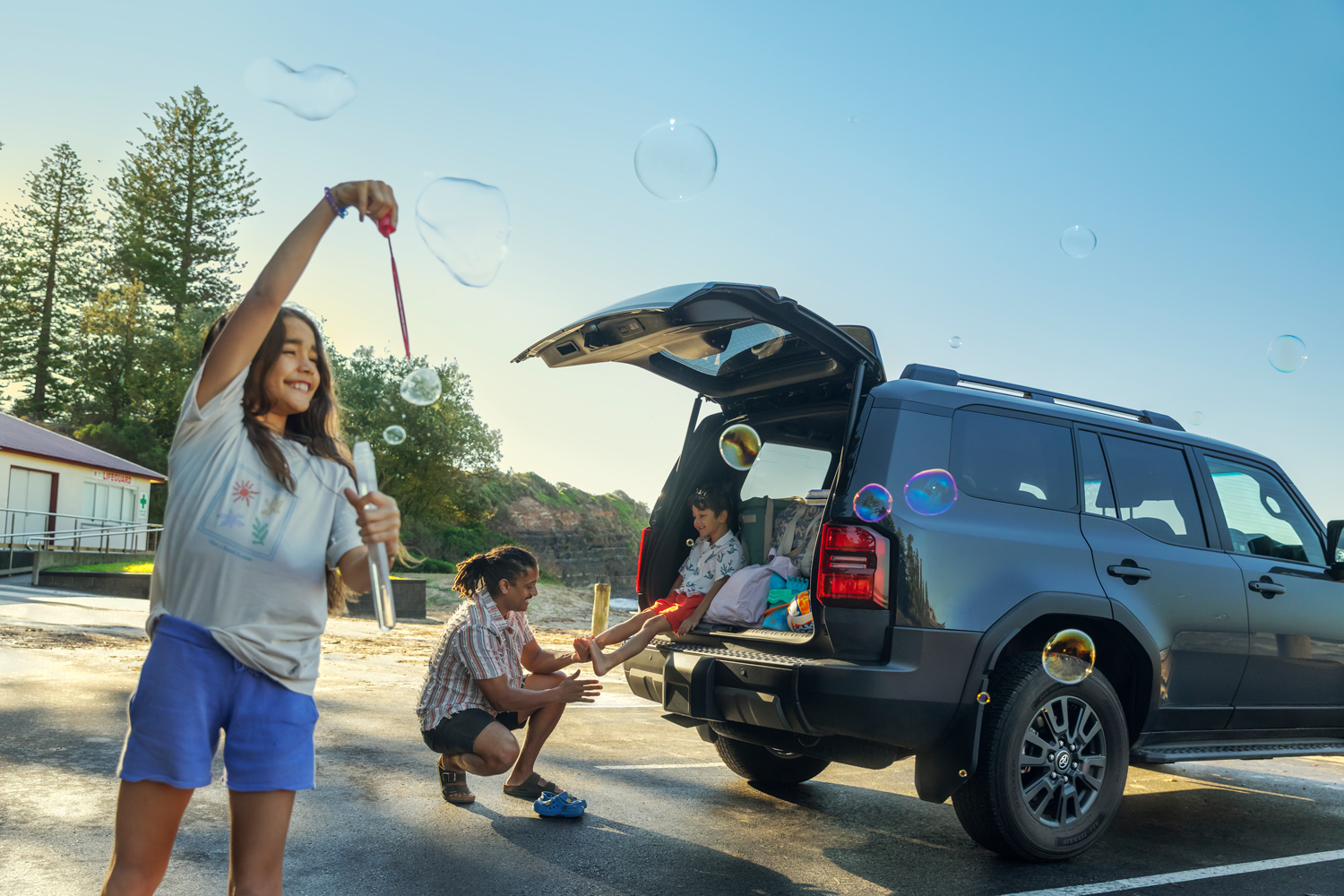 Young girl blowing soap bubbles in an outdoor carpark beside an open SUV tailgate with family members in the background.