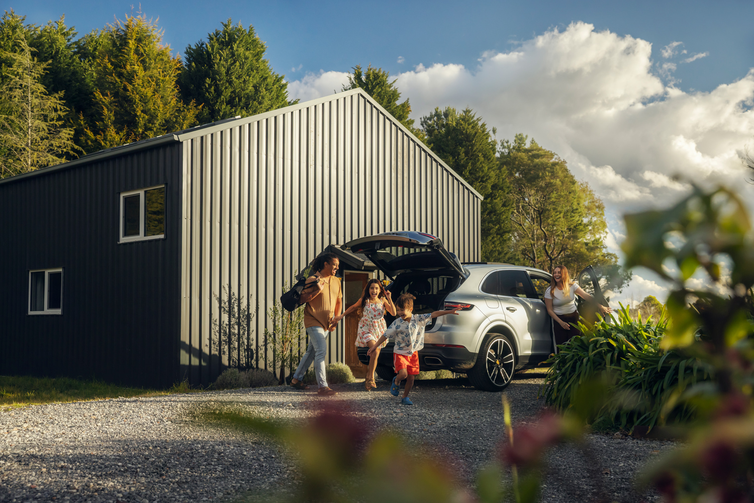 Family unpacking a silver SUV parked outside a dark metal building with open trunk and children running on a gravel driveway