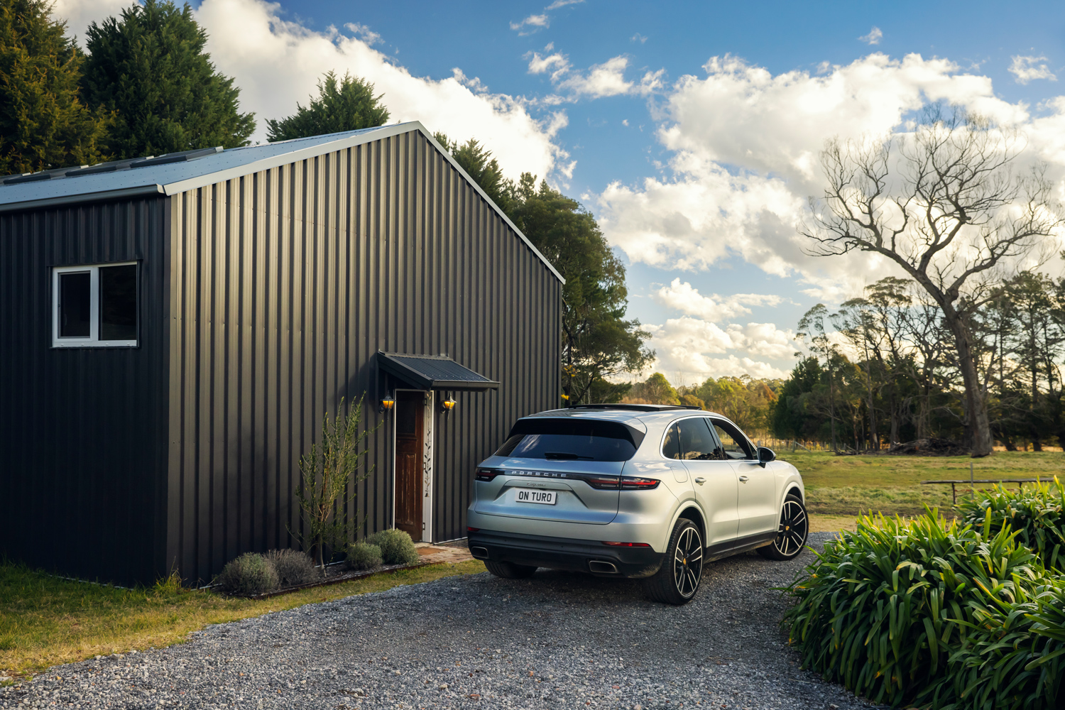 Silver Porsche SUV parked on a gravel driveway beside a modern dark corrugated metal barn in a rural countryside setting.