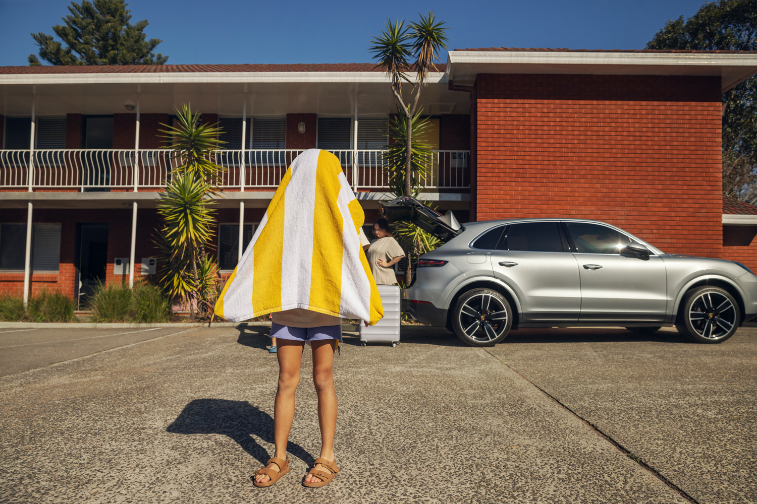 Child standing in a motel parking lot with a yellow and white striped towel draped over their head beside a silver SUV.
