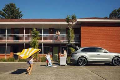 A family arrives at a brick motel with luggage and beach towels beside a parked silver SUV with an open trunk.
