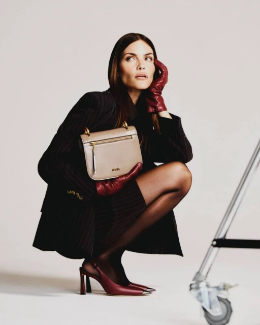 Woman in pinstripe suit and gloves poses with taupe handbag against white backdrop, with a silver lighting stand to the side.