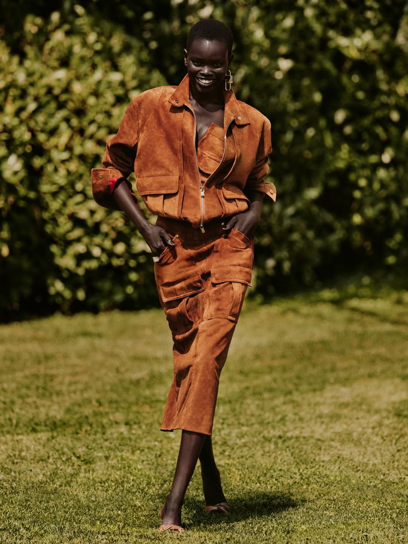 Athiec Geng smiling while walking in a brown suede jacket and matching midi skirt on a green lawn.