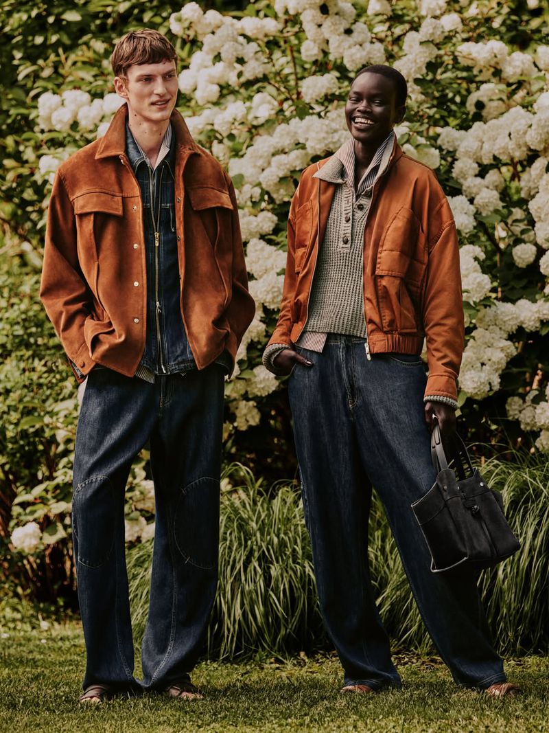 Two models standing outdoors against a backdrop of white hydrangeas wearing matching rust brown jackets and dark denim jeans