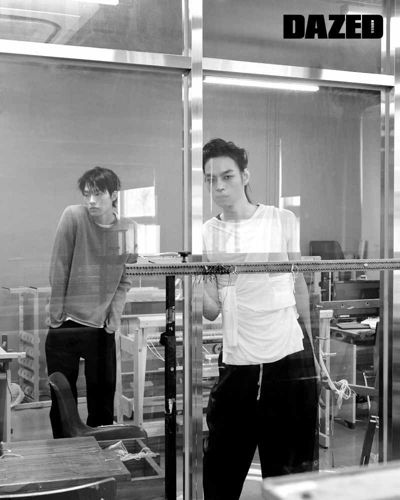 Monochrome fashion shot of two adult men indoors, reflected in a glass partition, wearing casual clothes in a studio setting.