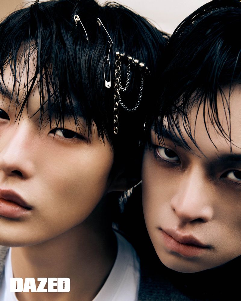 Close-up portrait of two young adult male models posing indoors with dark hair, safety pins, and chains, with a magazine logo.