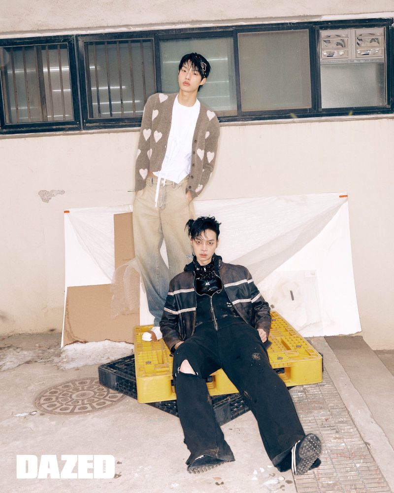 Two young men in stylish streetwear pose in an urban alley, one sitting on yellow pallets, the other standing near a boarded-up window.
