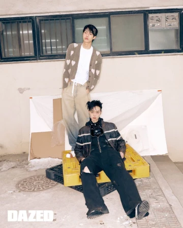 Two young men in stylish streetwear pose in an urban alley, one sitting on yellow pallets, the other standing near a boarded-up window.