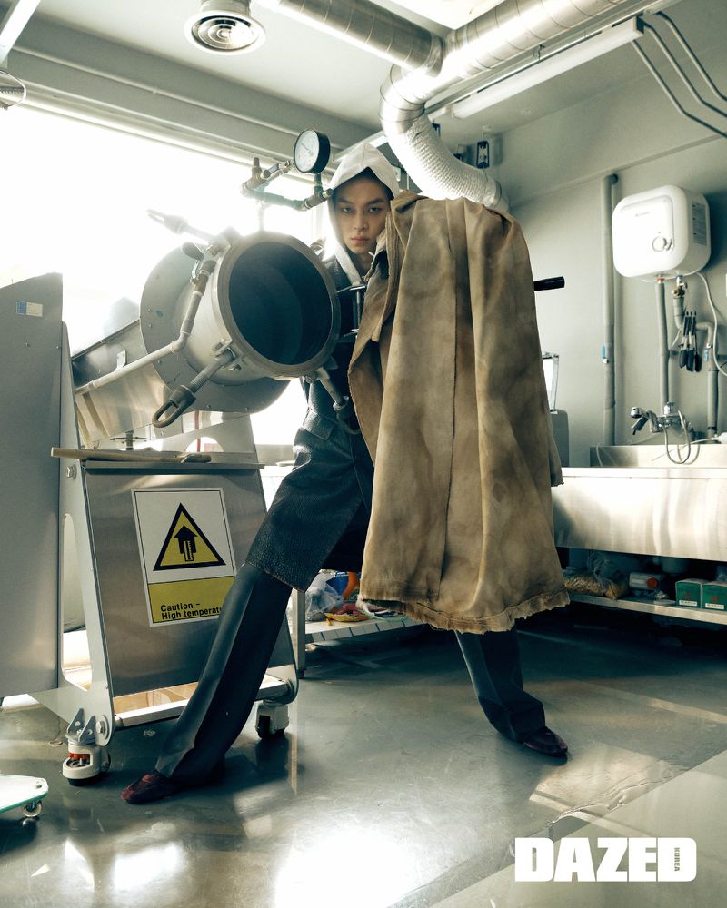 Adult standing inside industrial setting with a large metal machine, wearing dark jeans and jacket with tan jacket draped over shoulder.