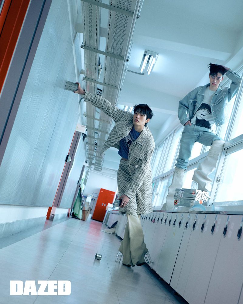 Two young adult male fashion models strike a pose in an indoor hallway setting with bright, cool-toned lighting and eye-level camera angle.