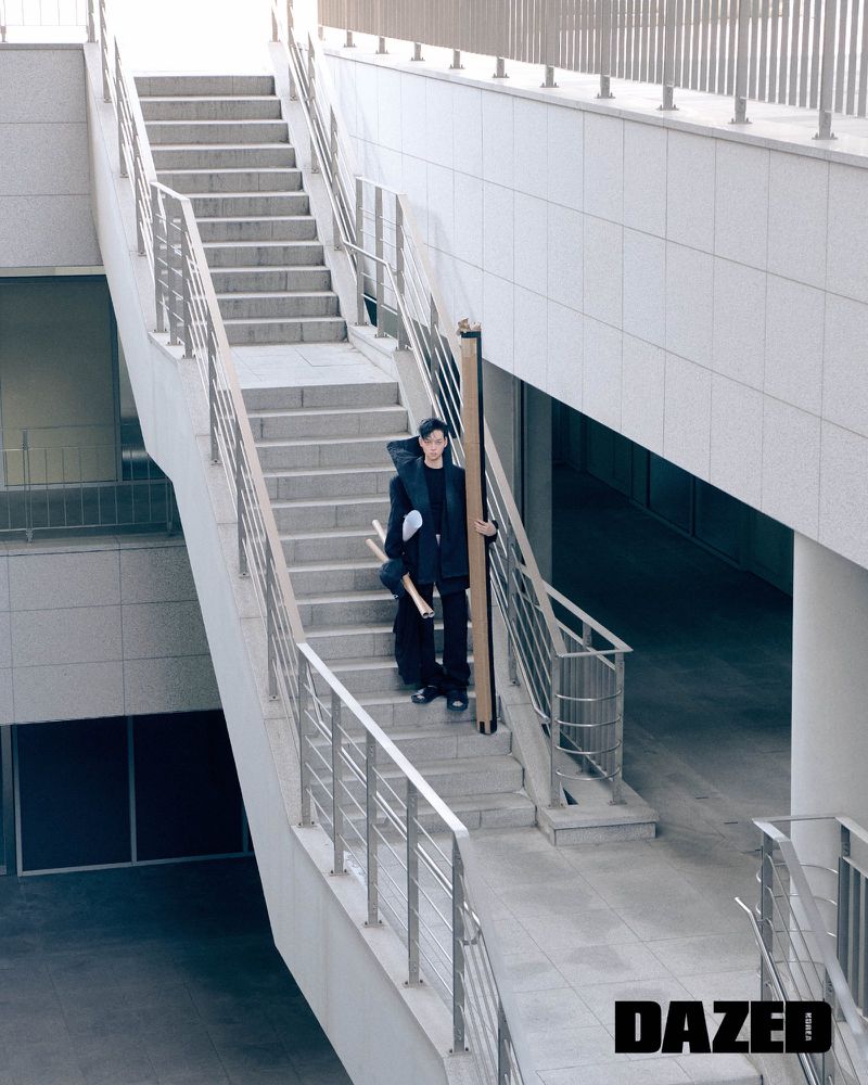 A stylish young adult male stands on outdoor stairs, wearing a black outfit and holding a long, rolled object, with a modern building in the background.