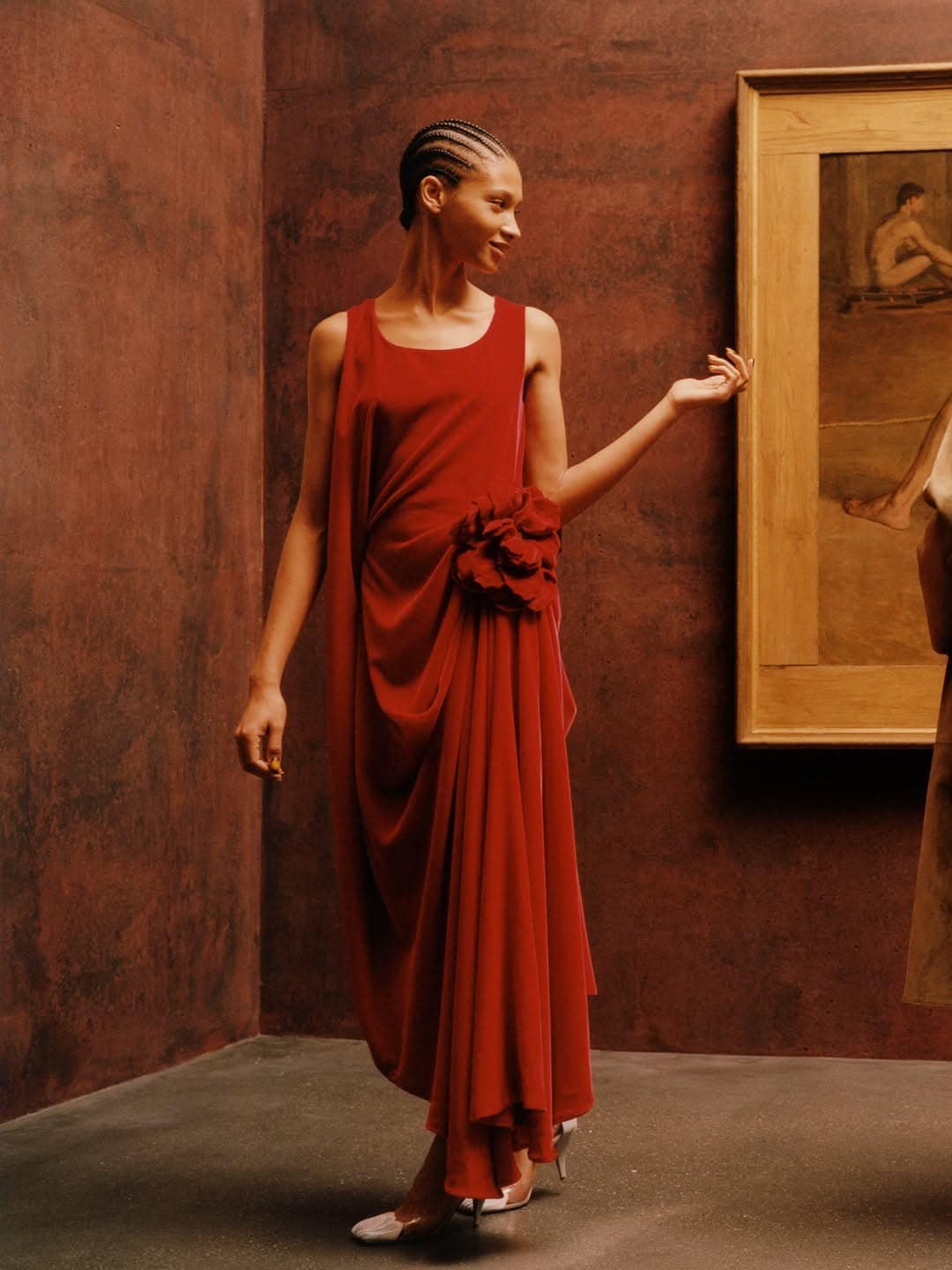 Model with cornrows wearing a draped red sleeveless gown with a floral rosette detail standing by a framed painting