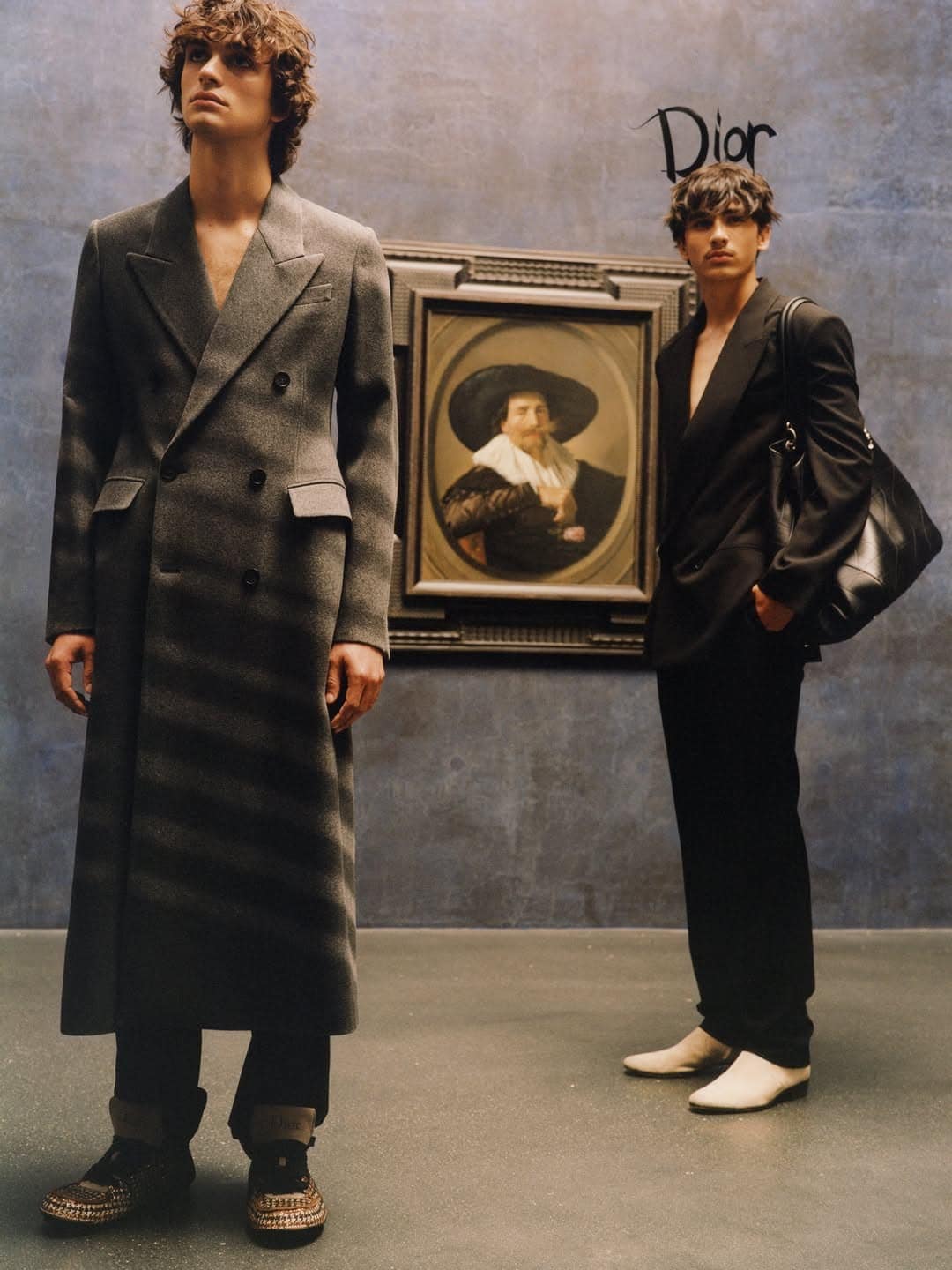 Two male models posing in tailored Dior menswear in front of a framed classical portrait in an art gallery.
