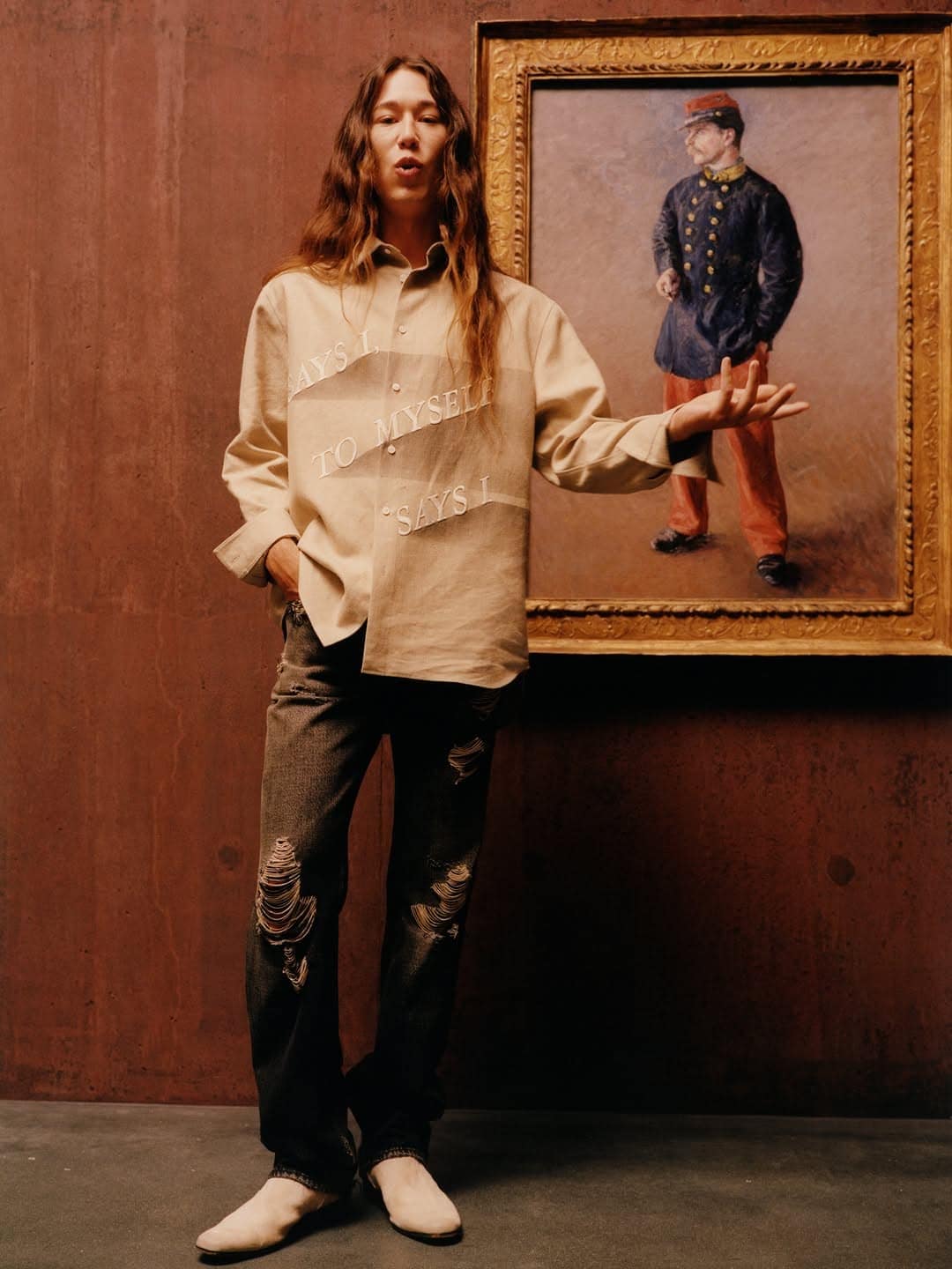 Model with long hair in a beige graphic shirt and distressed dark jeans gesturing toward a framed painting on a rust brown wall.