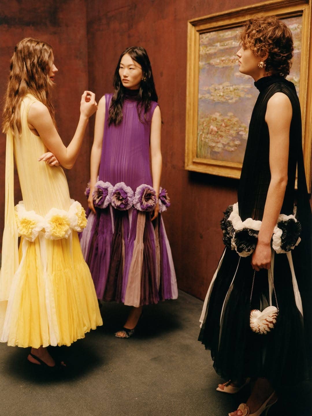 Three models wearing pleated dresses with flower trim standing in a gallery interior with a framed landscape painting on the wall