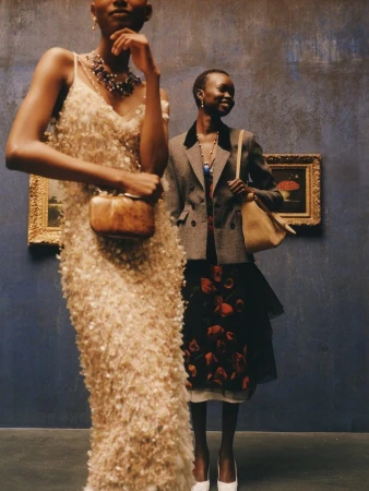 Two models in high fashion attire, one in a textured cream gown and another in a tailored blazer, posing against a blue gallery wall.