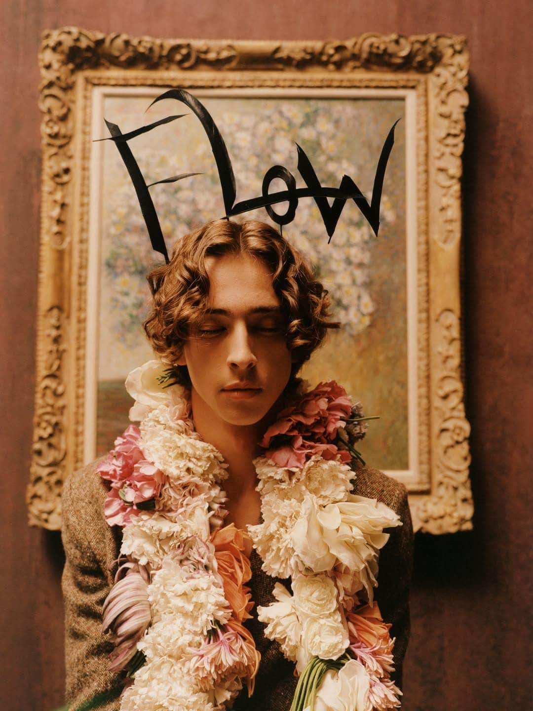 Man with curly brown hair wearing a heavy floral garland and tweed jacket before a framed oil painting with Flow lettering.