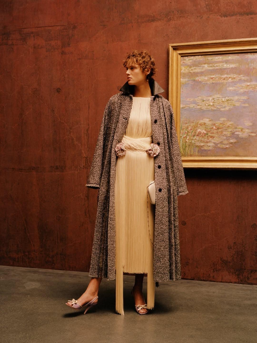 Model wearing a long tweed coat over a fringed cream dress posing beside a framed landscape painting in a rust-walled room.