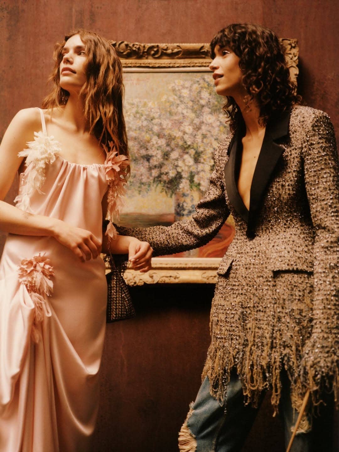 Two models pose before a framed floral painting in a blush pink satin dress and a fringed tweed blazer.