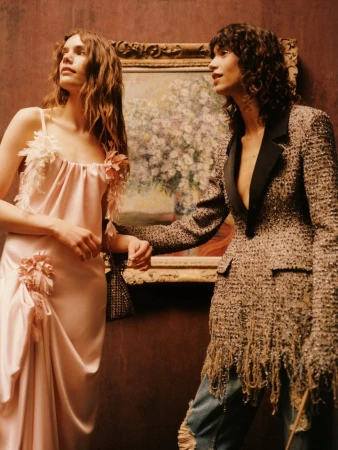 Two models pose before a framed floral painting in a blush pink satin dress and a fringed tweed blazer.