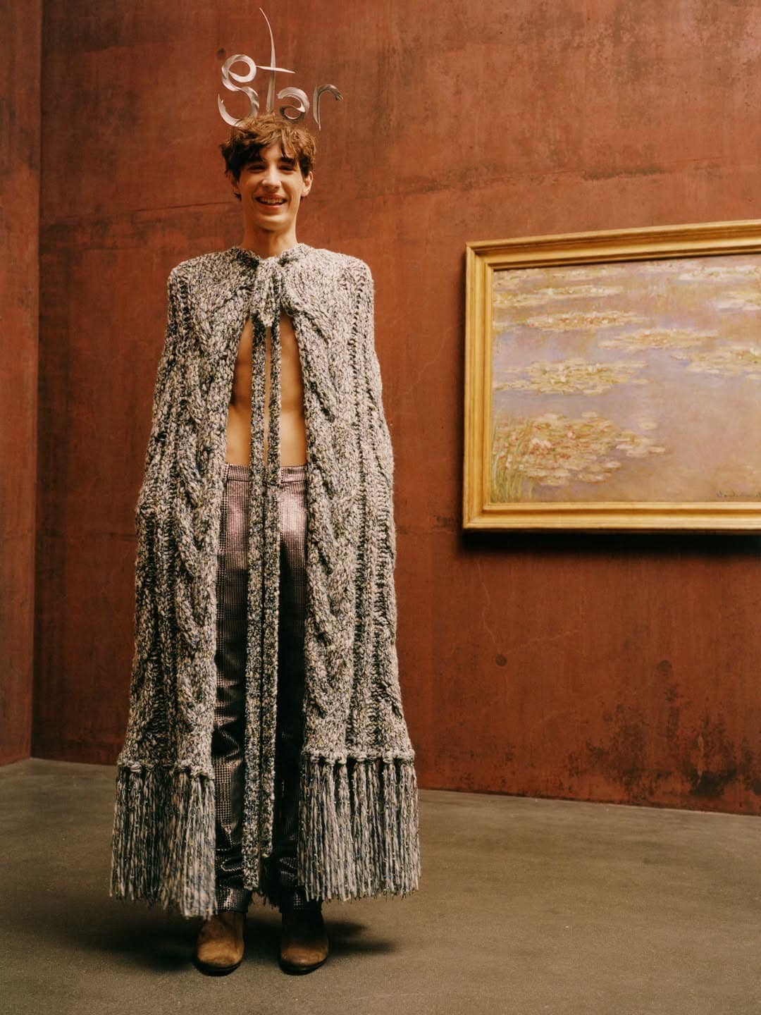 Smiling model in a floor-length cable knit fringe cape and wire star headpiece standing before a framed painting against a rust wall.