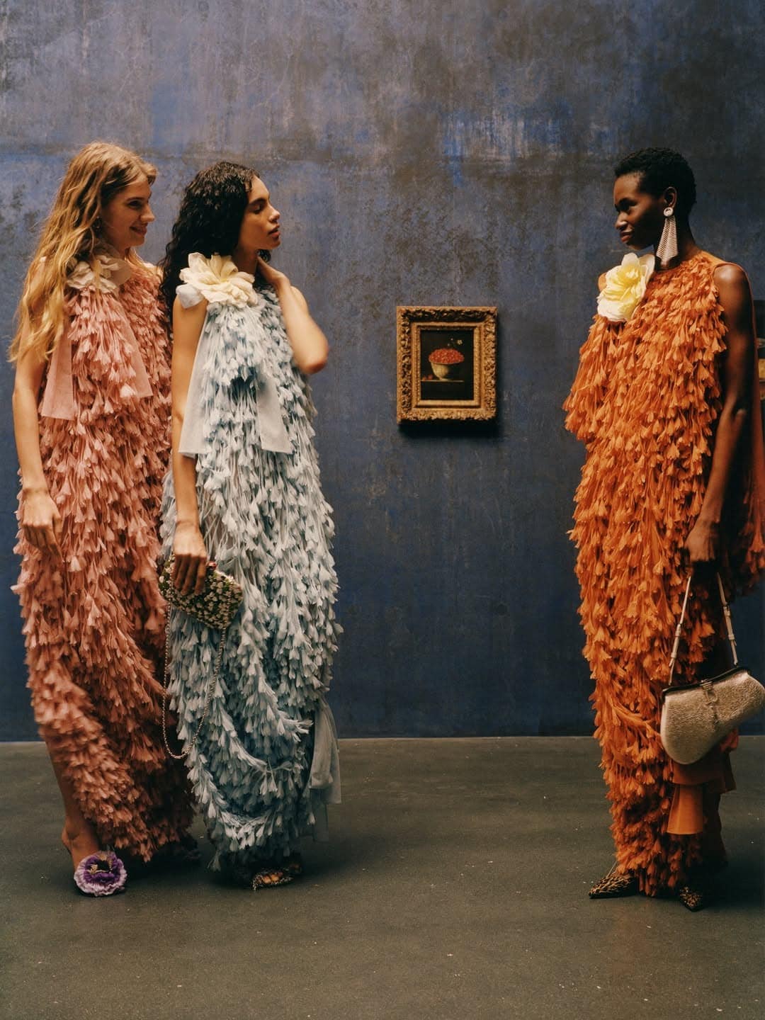 Three models wearing textured fringe dresses in pink, light blue, and orange standing before a blue wall with a framed painting.