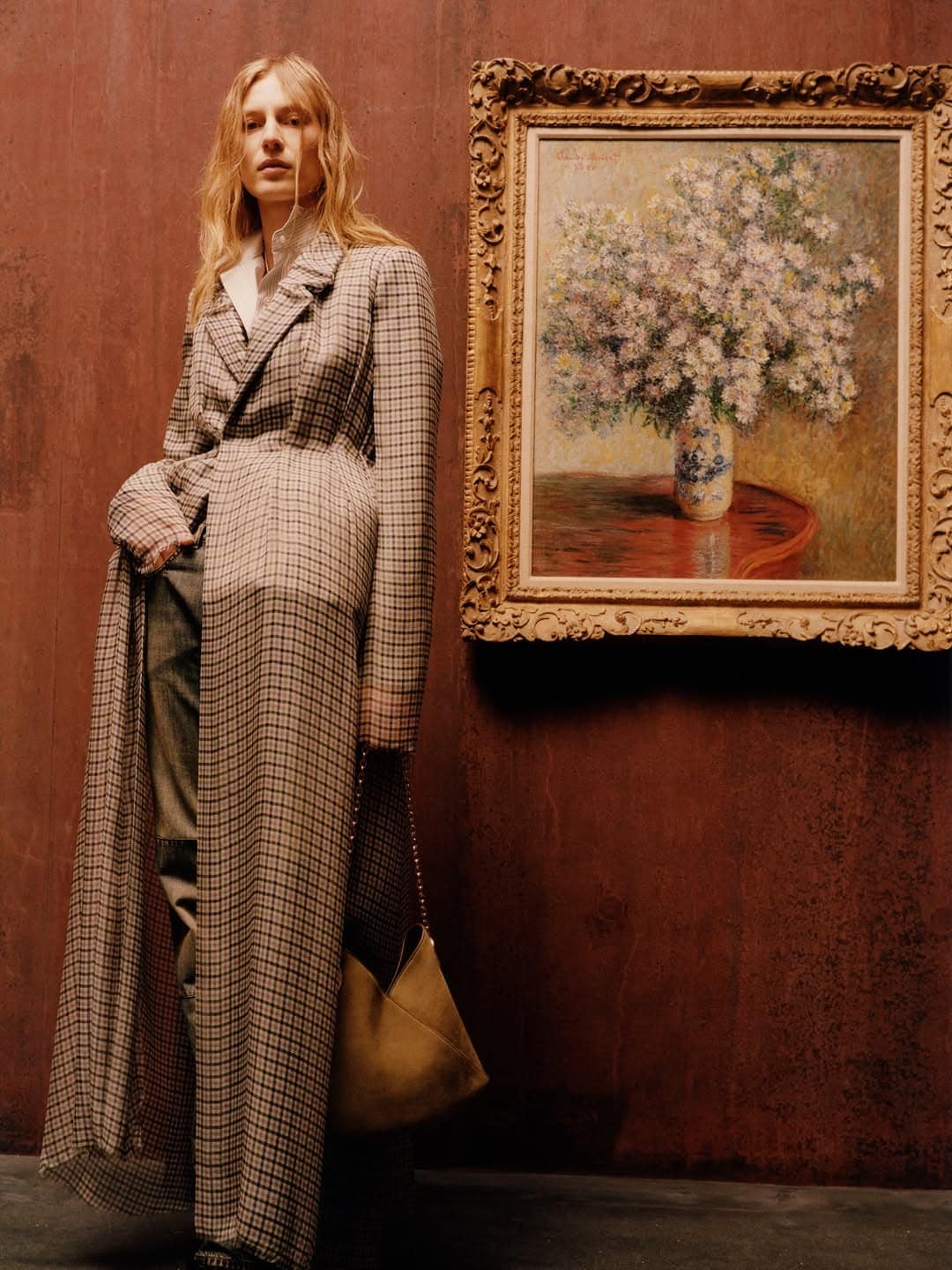Model wearing a floor-length plaid coat standing next to a framed floral oil painting against a textured rust-red studio wall.