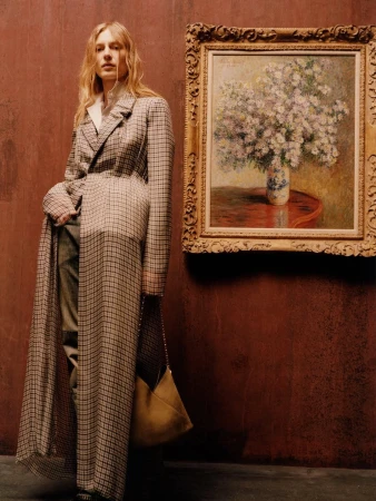 Model wearing a floor-length plaid coat standing next to a framed floral oil painting against a textured rust-red studio wall.