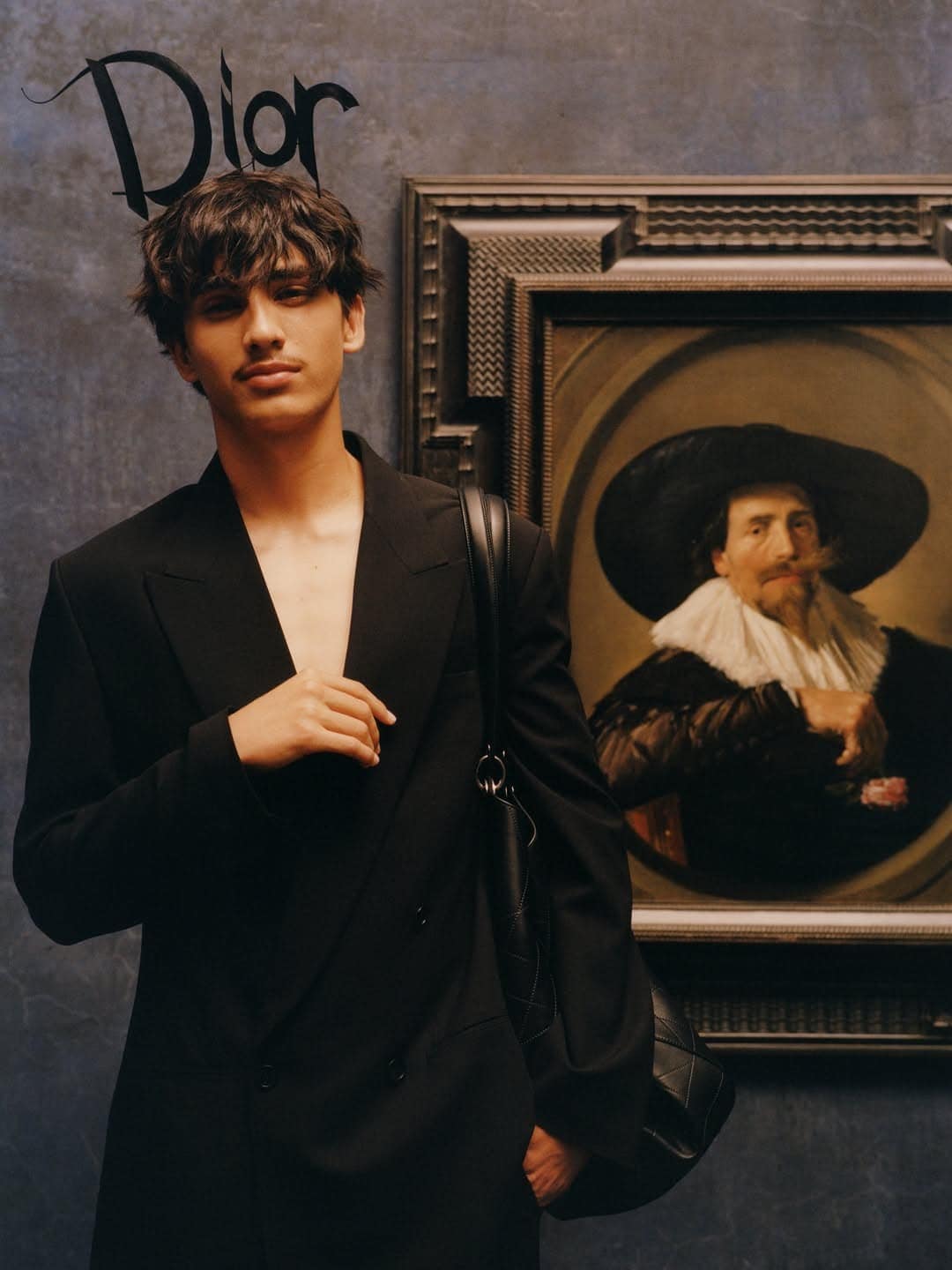 Model wearing a black tailored blazer and carrying a black shoulder bag beside a framed classical portrait painting.