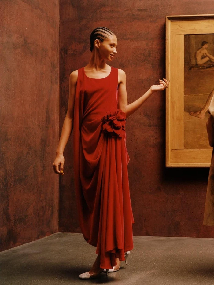 Model with cornrows wearing a draped red sleeveless gown with a floral rosette detail standing by a framed painting