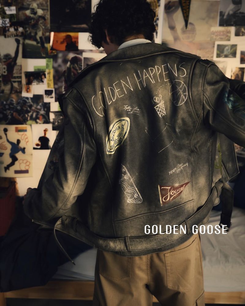 Rear view of a model wearing a customized distressed grey leather jacket with illustrations and patches in a poster-filled room.